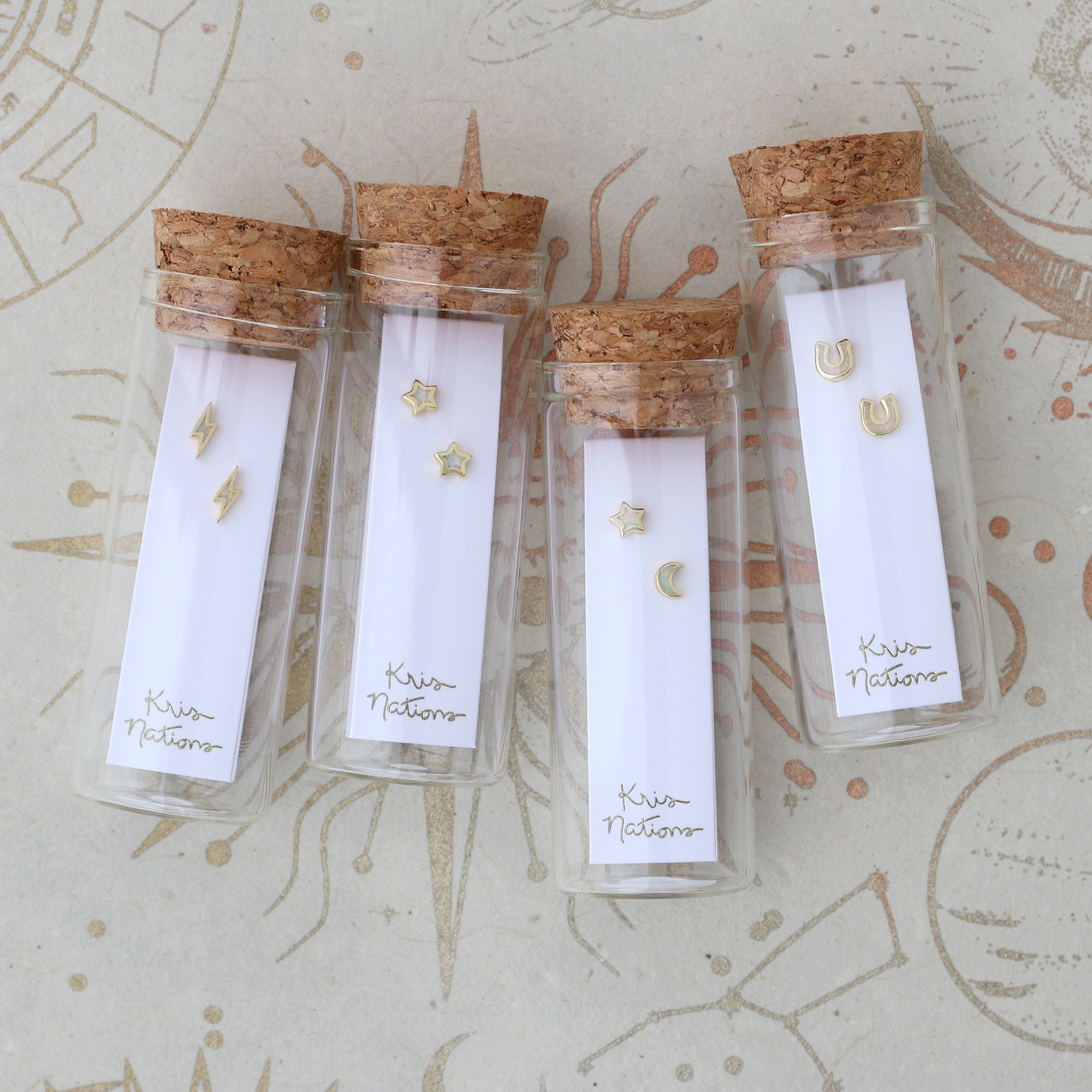 Four small glass bottles with cork lids containing gold earrings on a decorative background